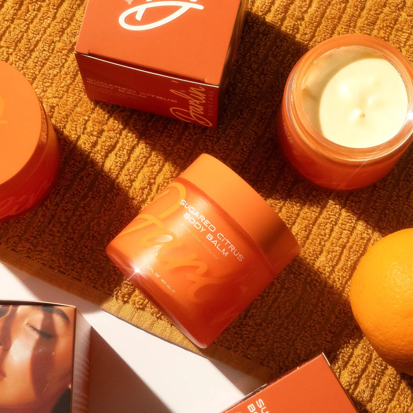 Orange body balm jar, candle, and orange fruit on a textured surface with a box in the background.