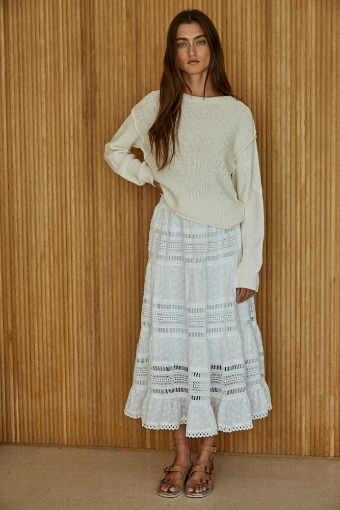 Woman wearing a white sweater and white lace skirt against a wooden paneled wall.