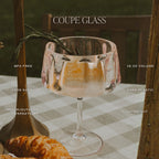 Coupe glass with a drink on a checkered tablecloth, surrounded by croissants and a wooden spoon.