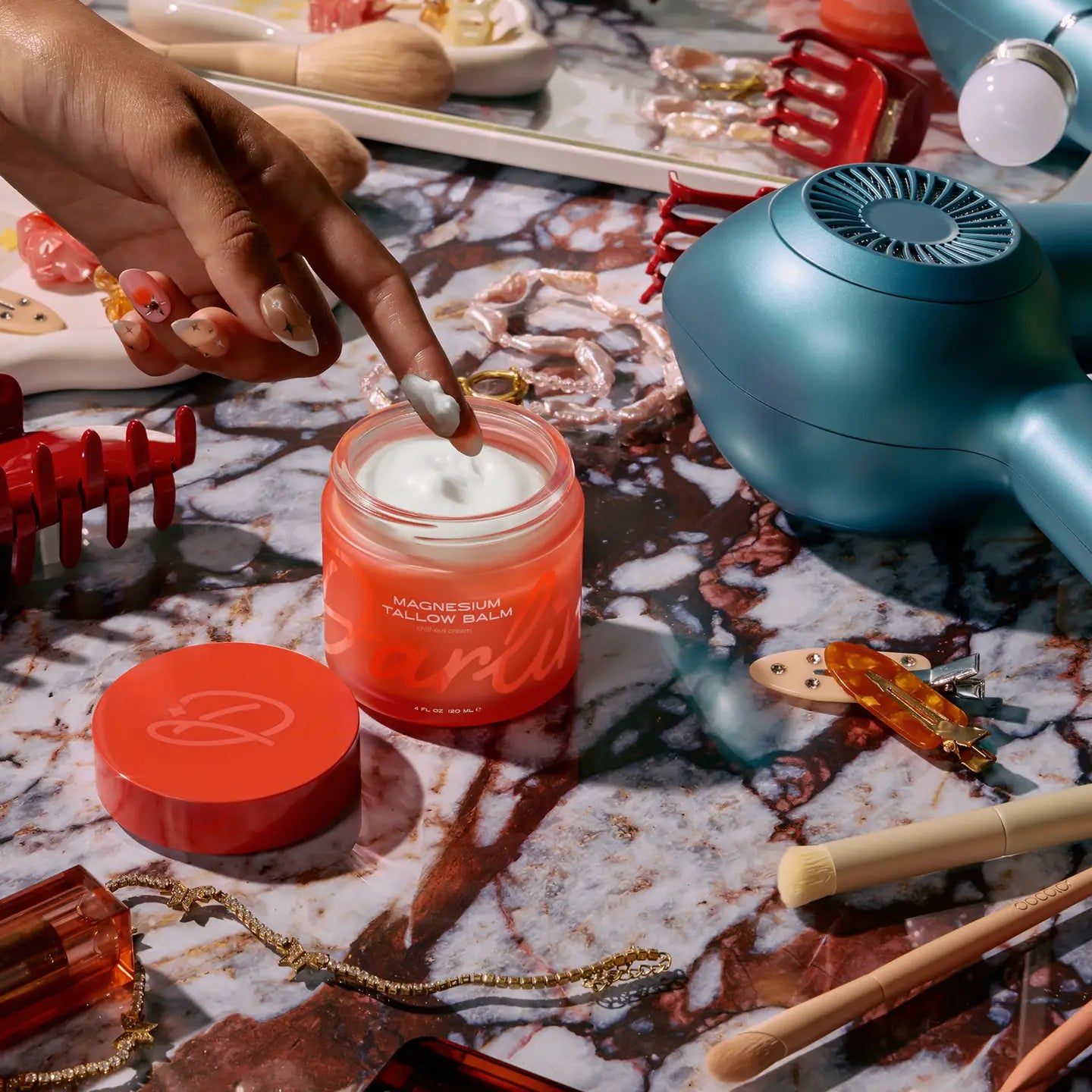 Person applying cream to a jar on a marble surface with beauty tools around