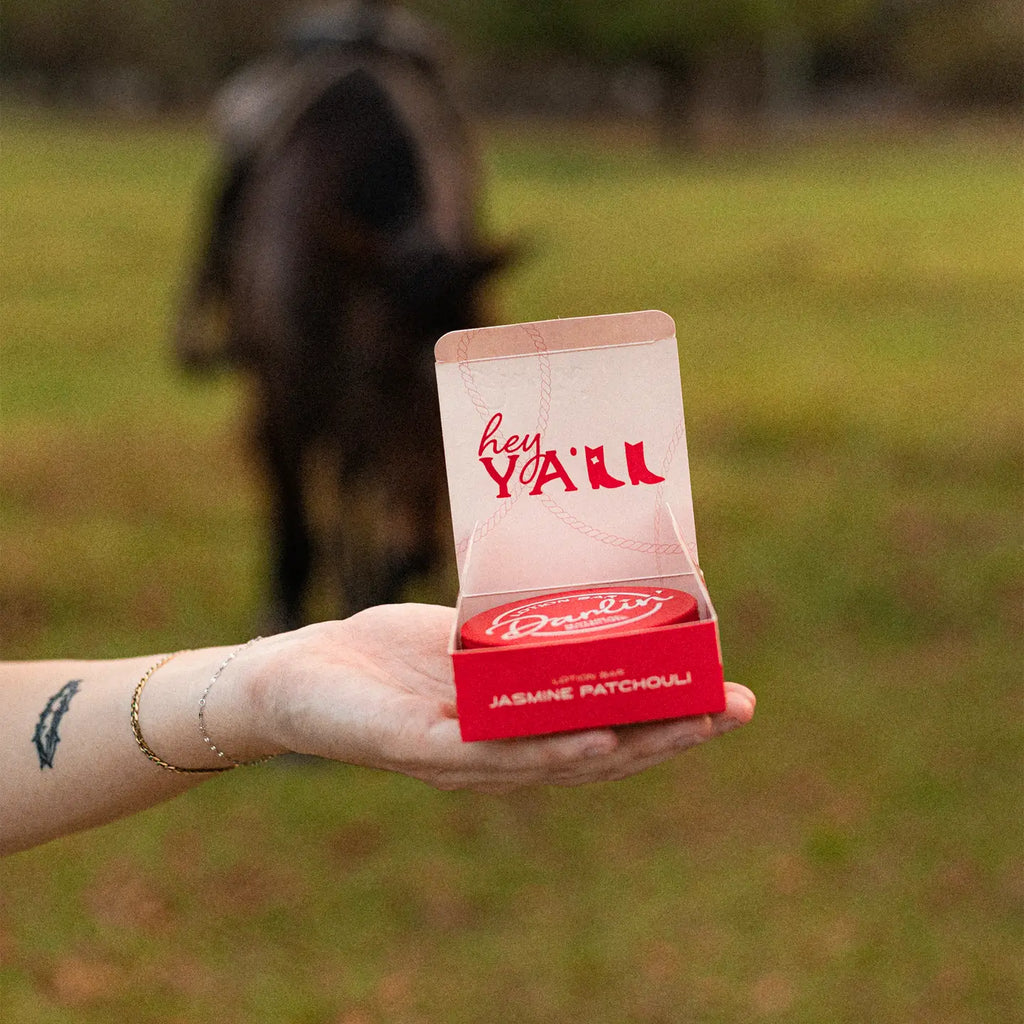 Hand holding a red and white product box with 'hey Y'all' text, blurred background of a person and horse.