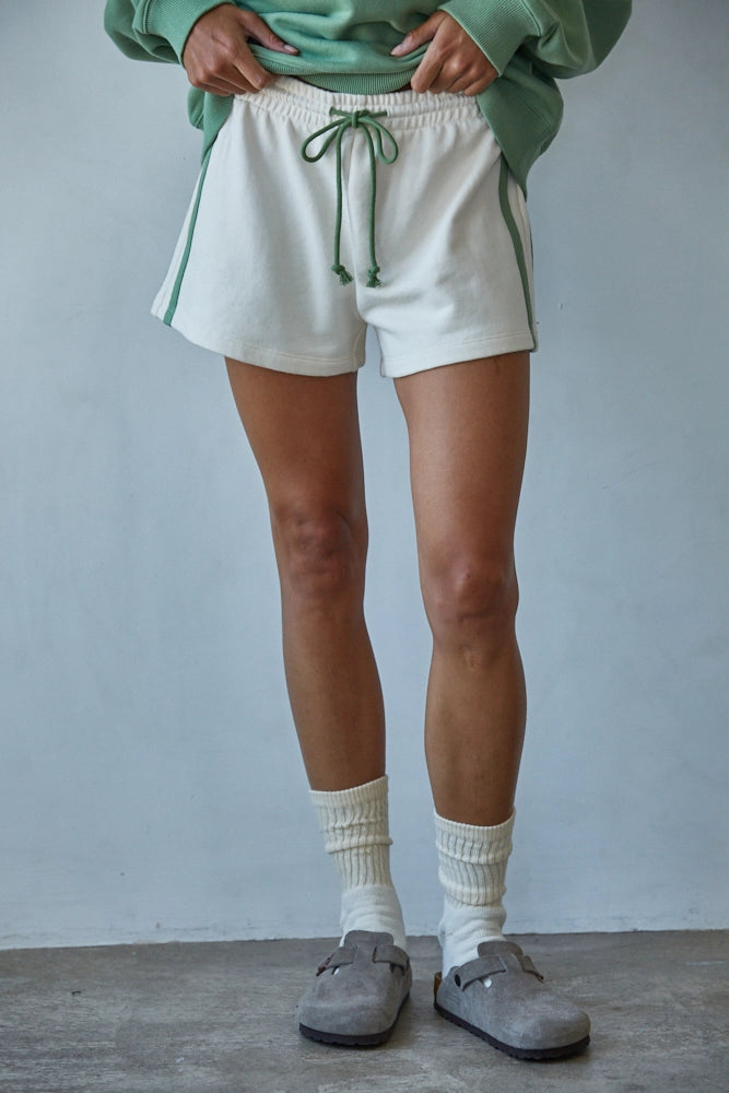 Person wearing white shorts, green sweater, and gray shoes against a plain background