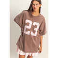 Woman wearing a brown oversized t-shirt with the number 23 printed on it against a plain background